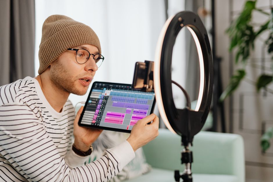 A young man wearing glasses and a beanie records a video indoors using a tablet and ring light.