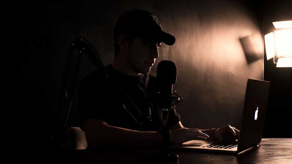 A man wearing a cap works on a laptop with a microphone setup in a dimly lit room.