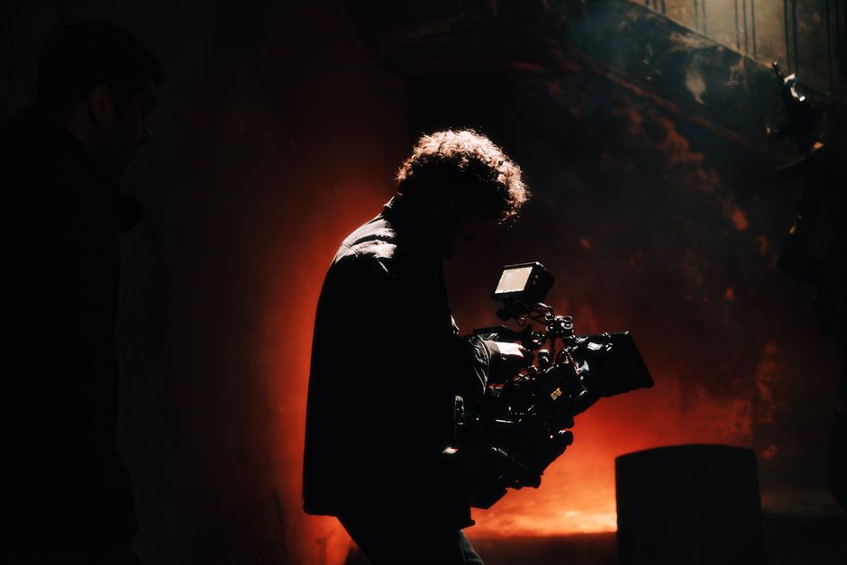 A professional cameraman captures footage in a creatively lit, moody setting, backlit by warm light.