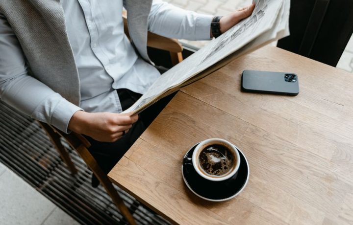 An adult male enjoying his coffee while reading a newspaper at a cafe with his smartphone nearby.