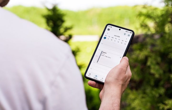 A person using a sauna booking app on a smartphone outdoors, surrounded by greenery.