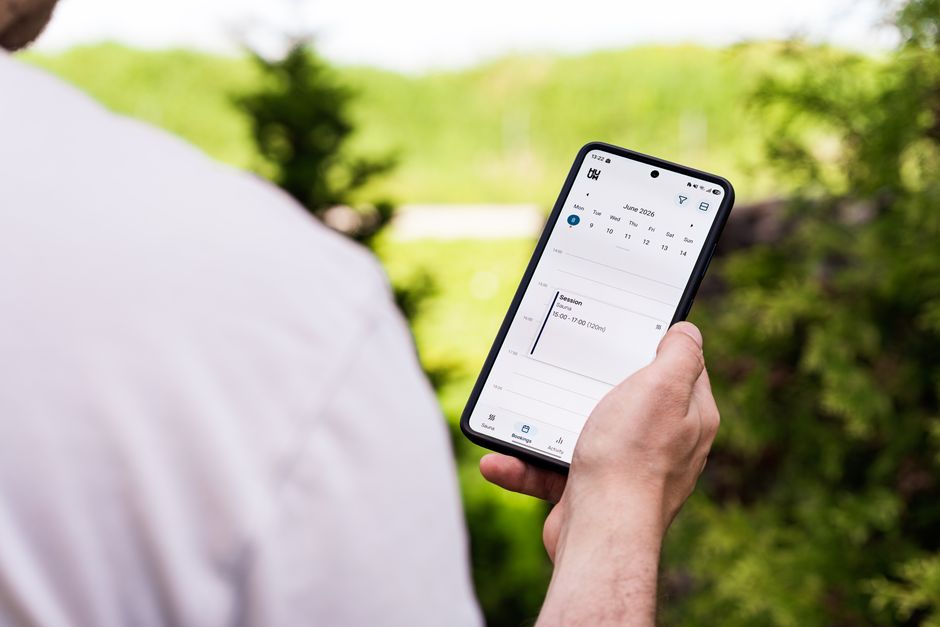 A person using a sauna booking app on a smartphone outdoors, surrounded by greenery.