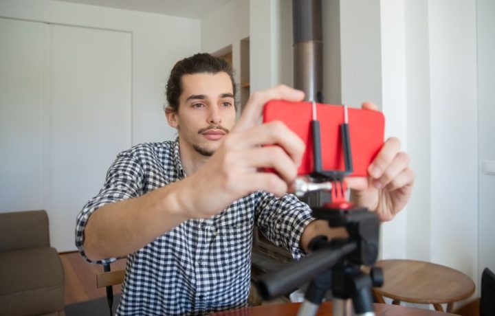 Confident young man adjusting smartphone on tripod for home vlogging setup.
