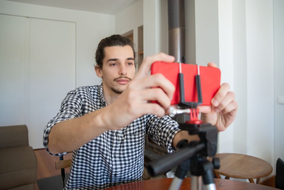 Confident young man adjusting smartphone on tripod for home vlogging setup.