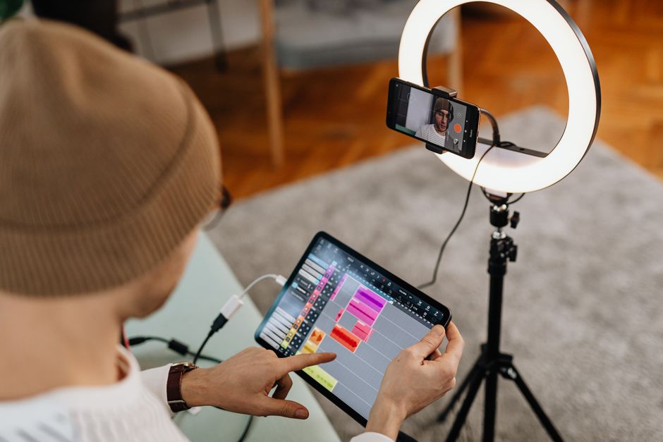 Man using a tablet with audio editing software in a home recording setup with a ring light and smartphone.