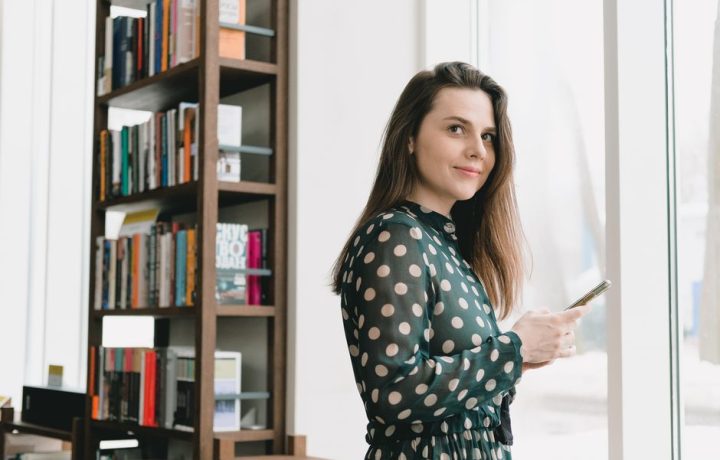 Side view of positive young female student with long dark hair in elegant dress using smartphone and smiling while standing near window in light library
