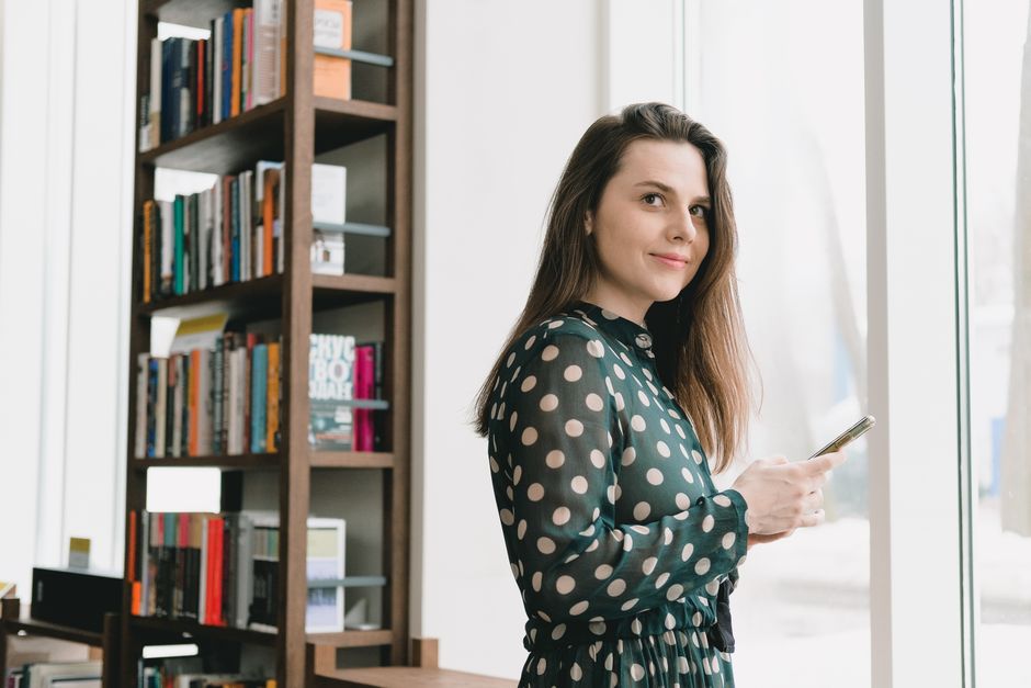 Side view of positive young female student with long dark hair in elegant dress using smartphone and smiling while standing near window in light library