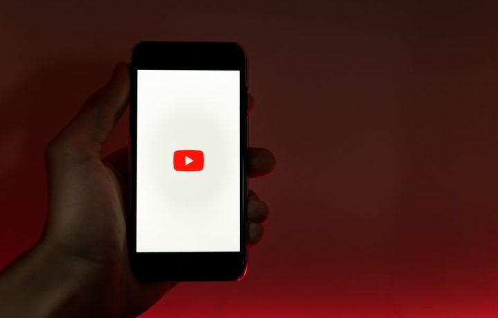 A hand holding a smartphone displaying the YouTube app against a red background.