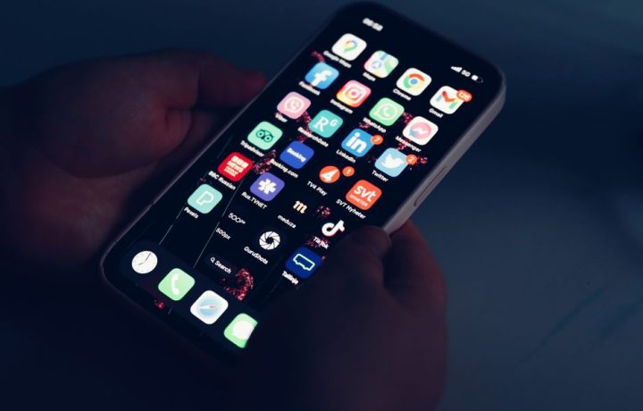 Close-up of hands holding a smartphone displaying various app icons in a dimly lit room.
