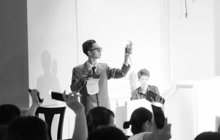 Elegant black and white photo of an auction scene with a presenter and bidders raising phones.