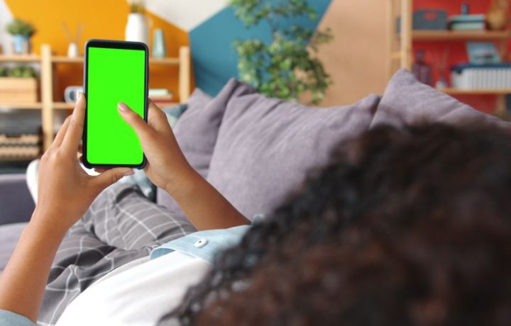 Woman lying on couch holding a green screen smartphone indoors, ideal for apps.