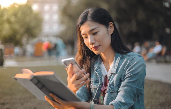 A young woman in a denim jacket sits outdoors reading a book and using her smartphone, enjoying a sunny day.