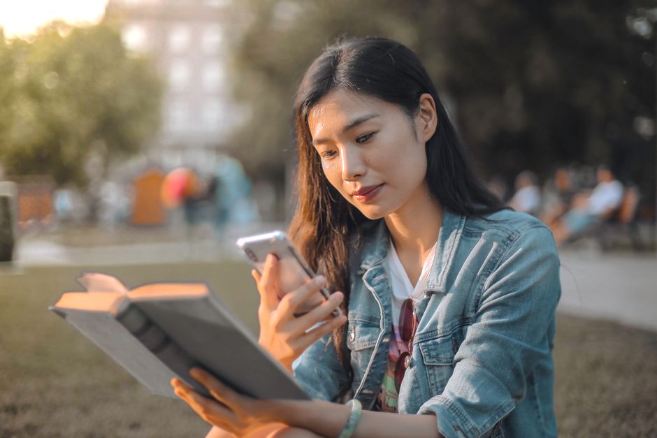 A young woman in a denim jacket sits outdoors reading a book and using her smartphone, enjoying a sunny day.