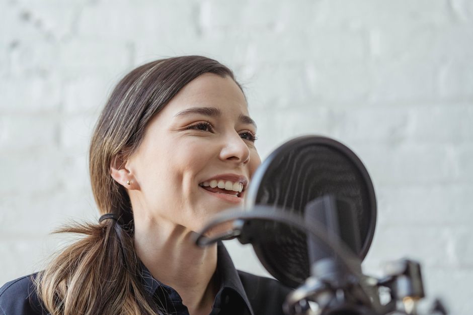 Positive female recording podcast on mic while working in light studio against white brick wall