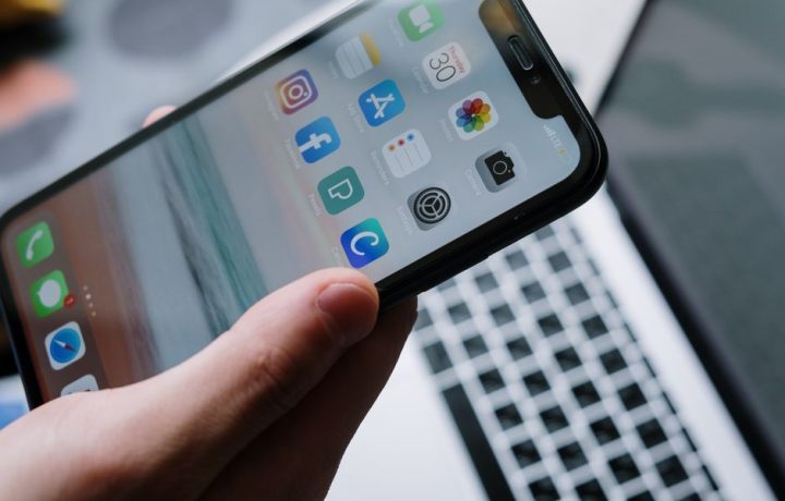 Person holding a smartphone in front of a laptop showcasing various app icons, highlighting digital connectivity.
