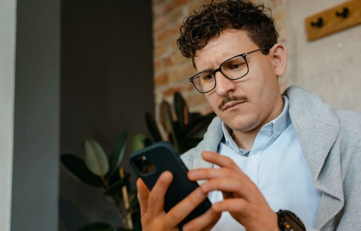 A focused man with glasses using a smartphone indoors next to a brick wall.