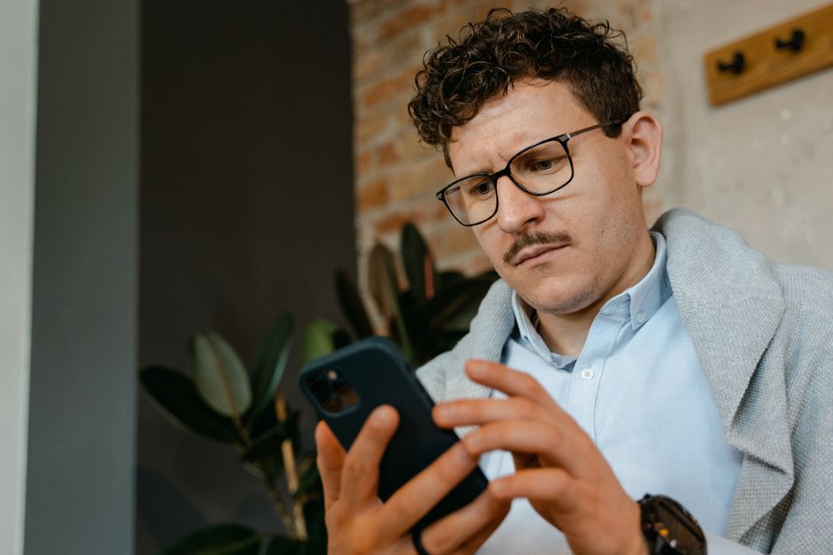 A focused man with glasses using a smartphone indoors next to a brick wall.