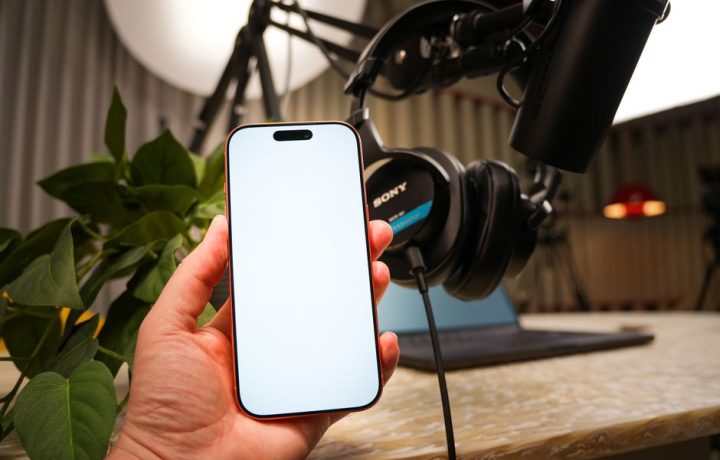 A hand holding a smartphone in a recording studio with microphone and headphones on a desk.