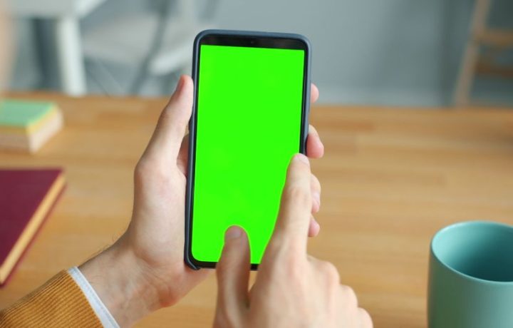 A person interacts with a smartphone displaying a green screen at a wooden desk with a mug.