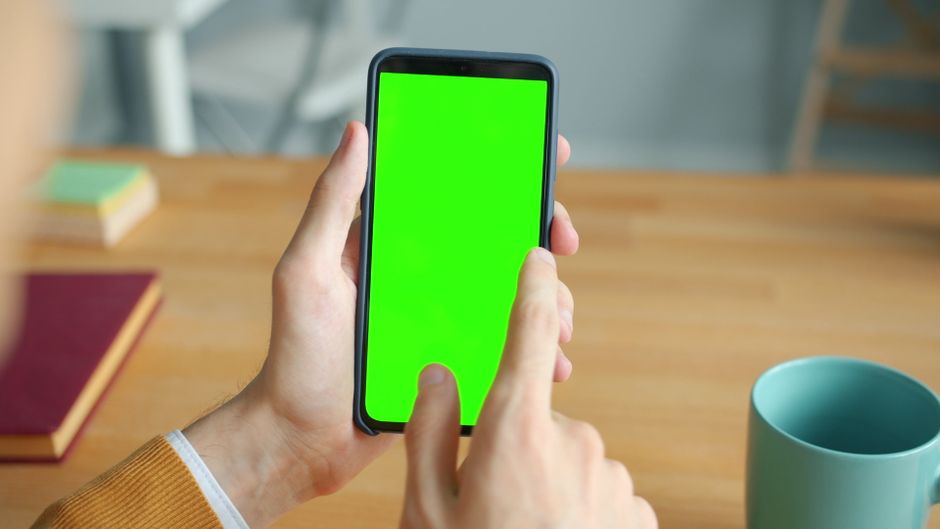 A person interacts with a smartphone displaying a green screen at a wooden desk with a mug.