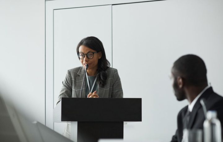 African American businesswoman delivering a speech at a conference podium with a colleague listening.