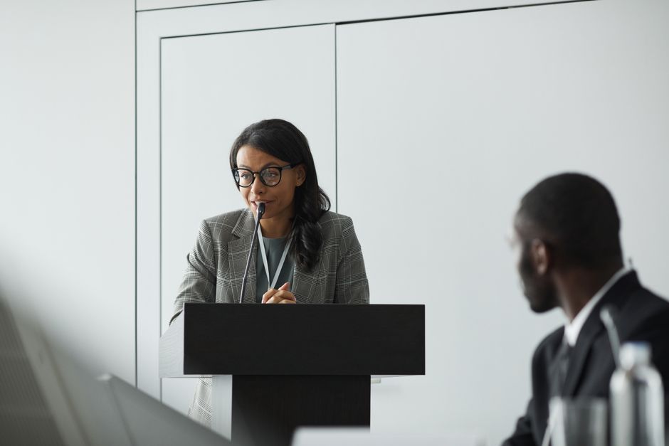 African American businesswoman delivering a speech at a conference podium with a colleague listening.