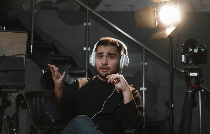 Portrait of a man wearing headphones, sitting in a studio, gesturing towards the camera, with a microphone and lighting setup.