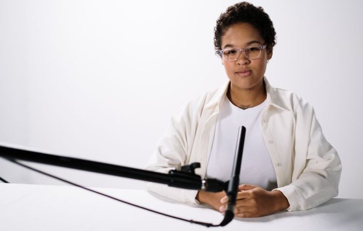 Portrait of woman in white shirt sitting at a table with a microphone in a studio setting.