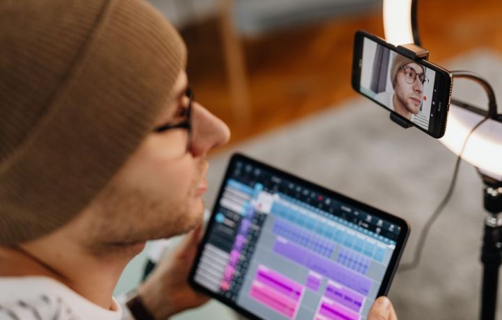 Man with eyeglasses and knitted cap using tablet for video call, illuminated by ring light.