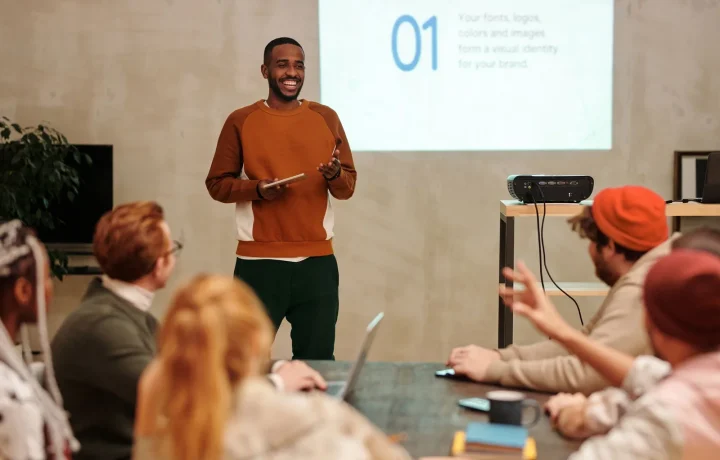 A man confidently presents in a sleek office setting to an engaged and diverse audience