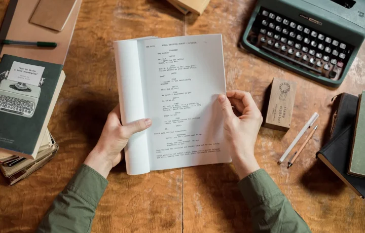 Hands holding a script page on a vintage desk with a typewriter and books, illustrating script writing for teleprompters
