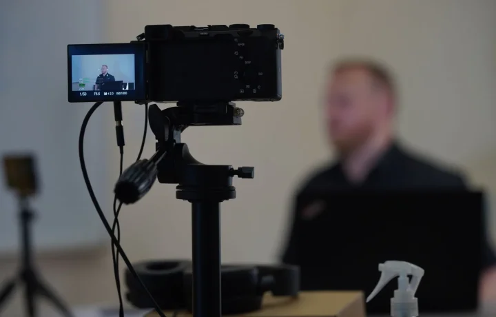 Close-up of a camera setup with a blurred speaker, illustrating the use of a teleprompter for video formats