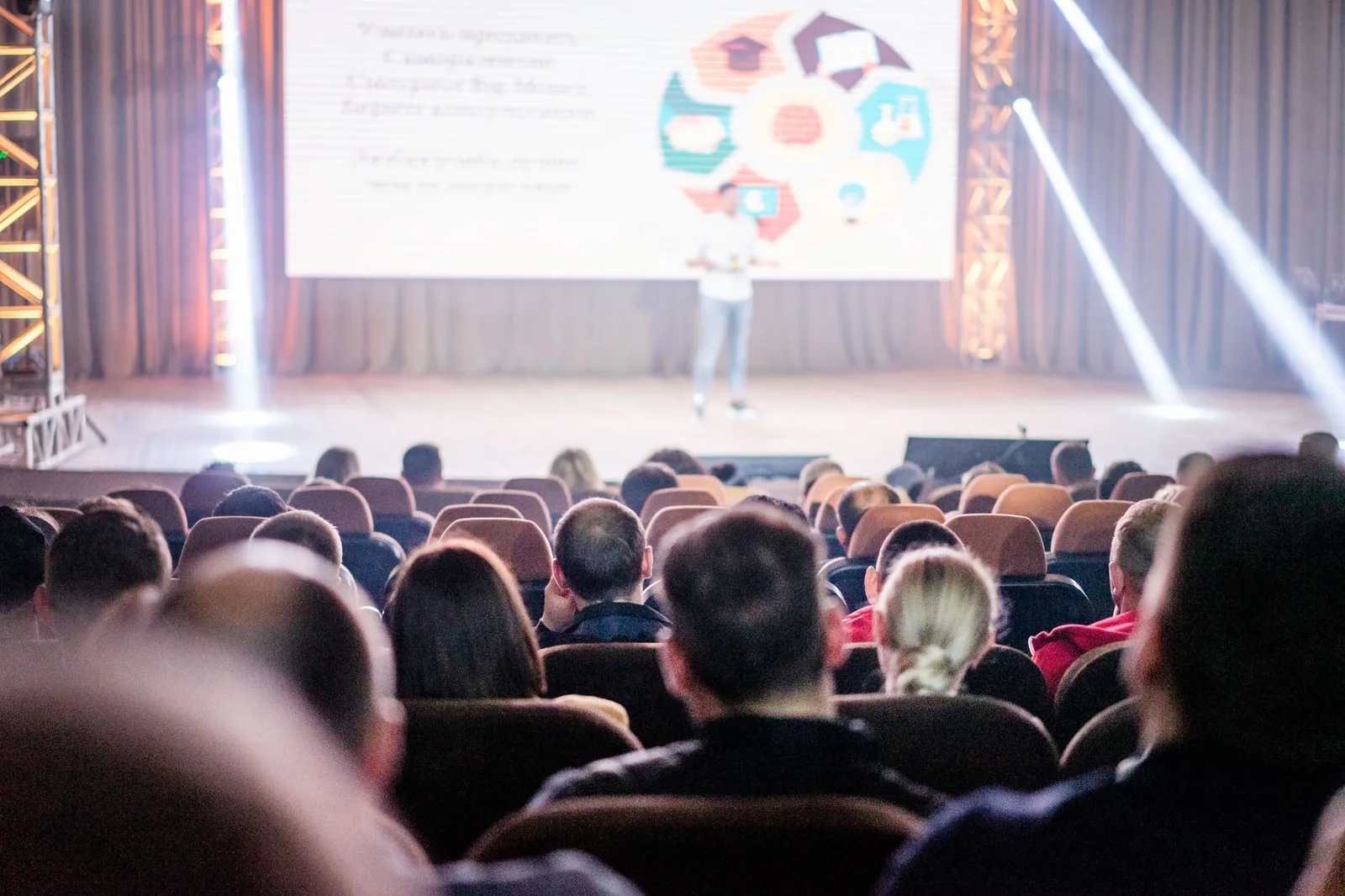 Conference attendees watching a speaker on a brightly lit stage during a live presentation